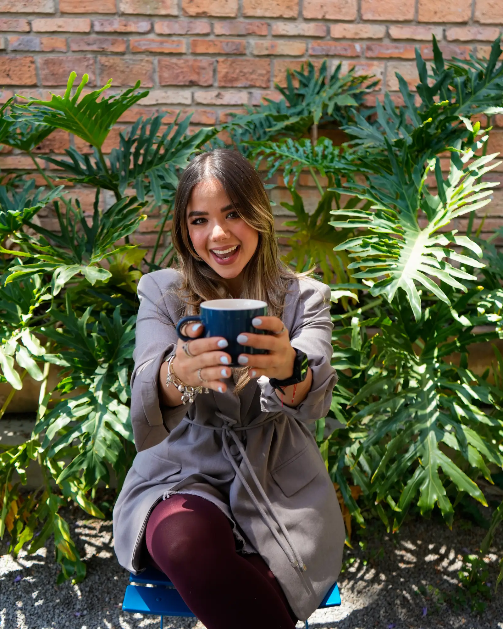 Clienta sonriente sosteniendo una taza de café frente a una pared de ladrillo con plantas